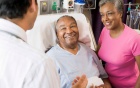 Man lying in hospital bed with woman at his side; both speaking with a physician. 